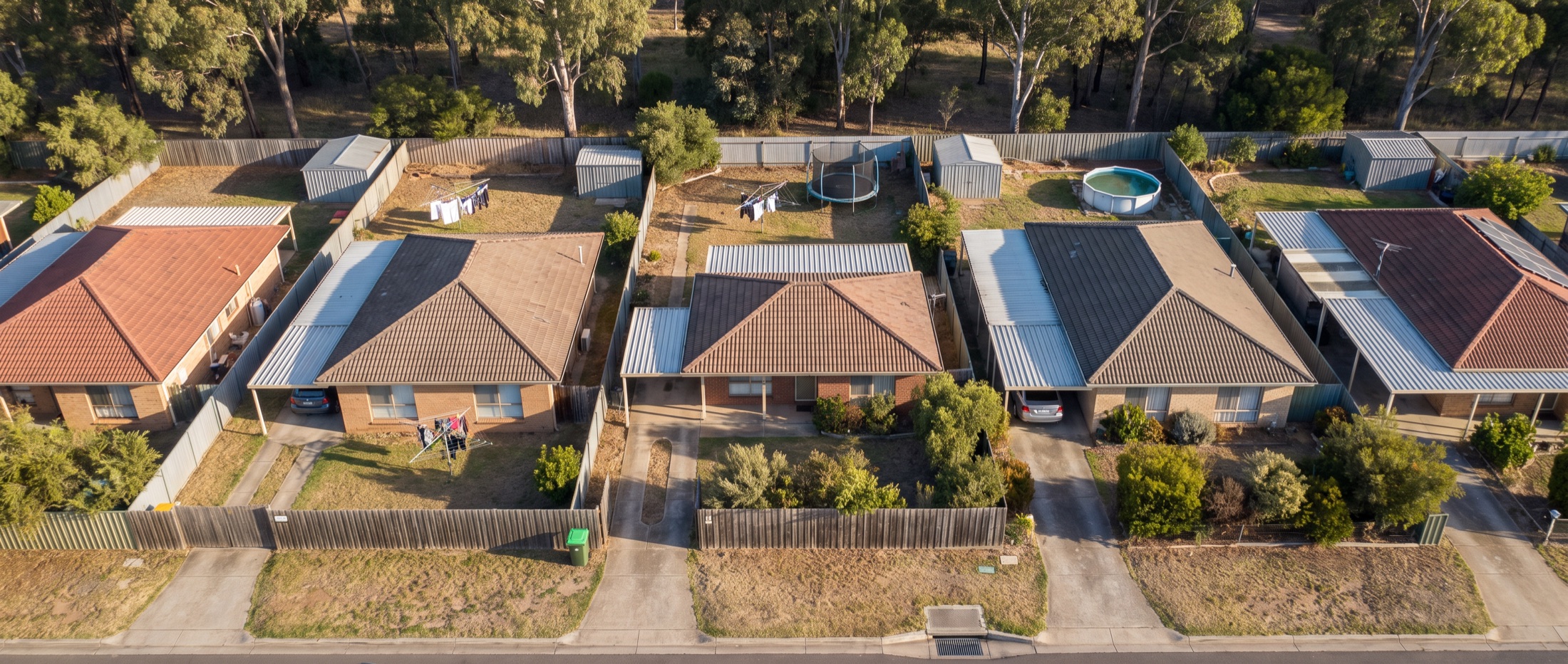 An ordinary Australian suburban street photographed from directly overhead, showing each house on its own block with fence lines, sheds, driveways and a stormwater grate at the kerb