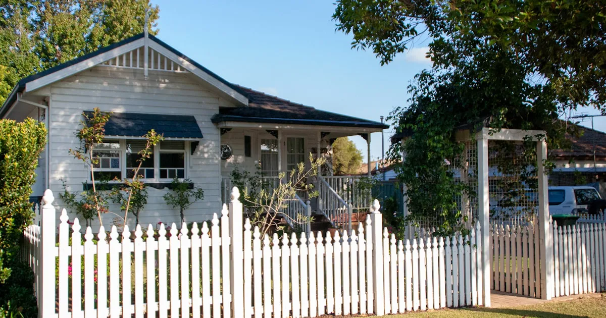 A single-storey weatherboard house behind a low white picket fence, with a leafy front garden