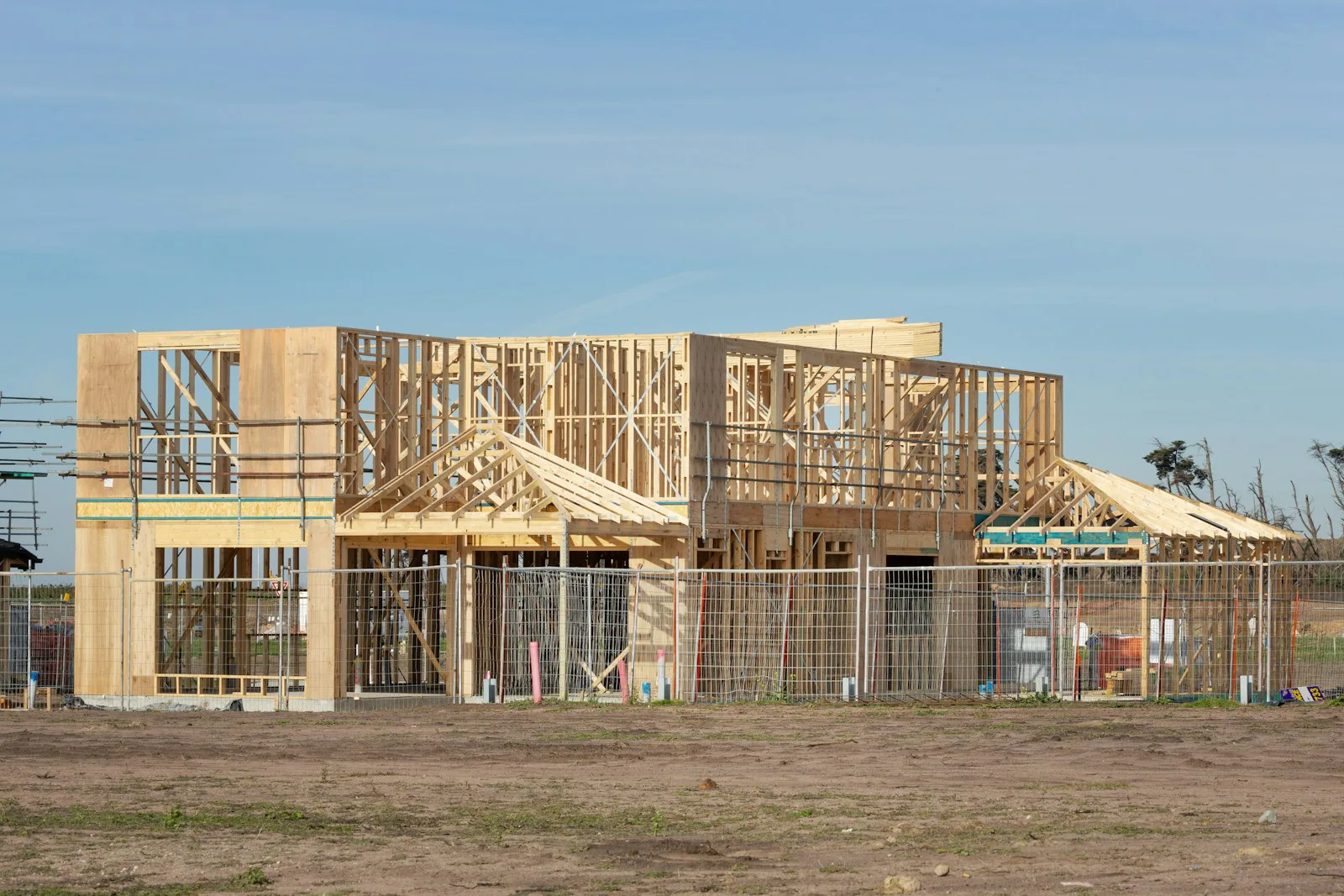 A two-storey timber house frame under construction behind temporary site fencing, with roof trusses stacked on the upper floor waiting to be lifted into place
