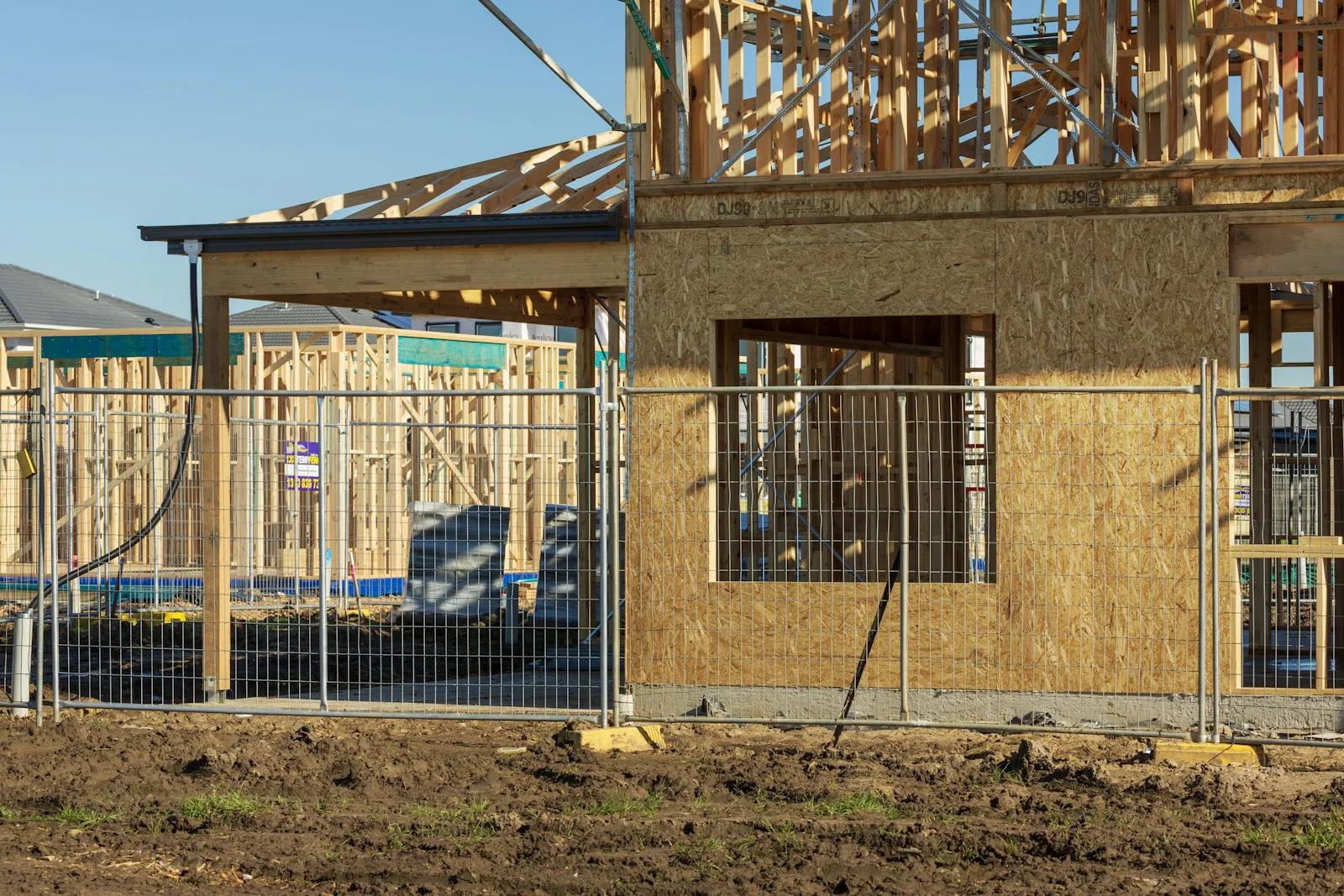 An unfinished two-storey timber house frame standing empty behind temporary mesh site fencing, its ground floor part-sheeted and no workers on site