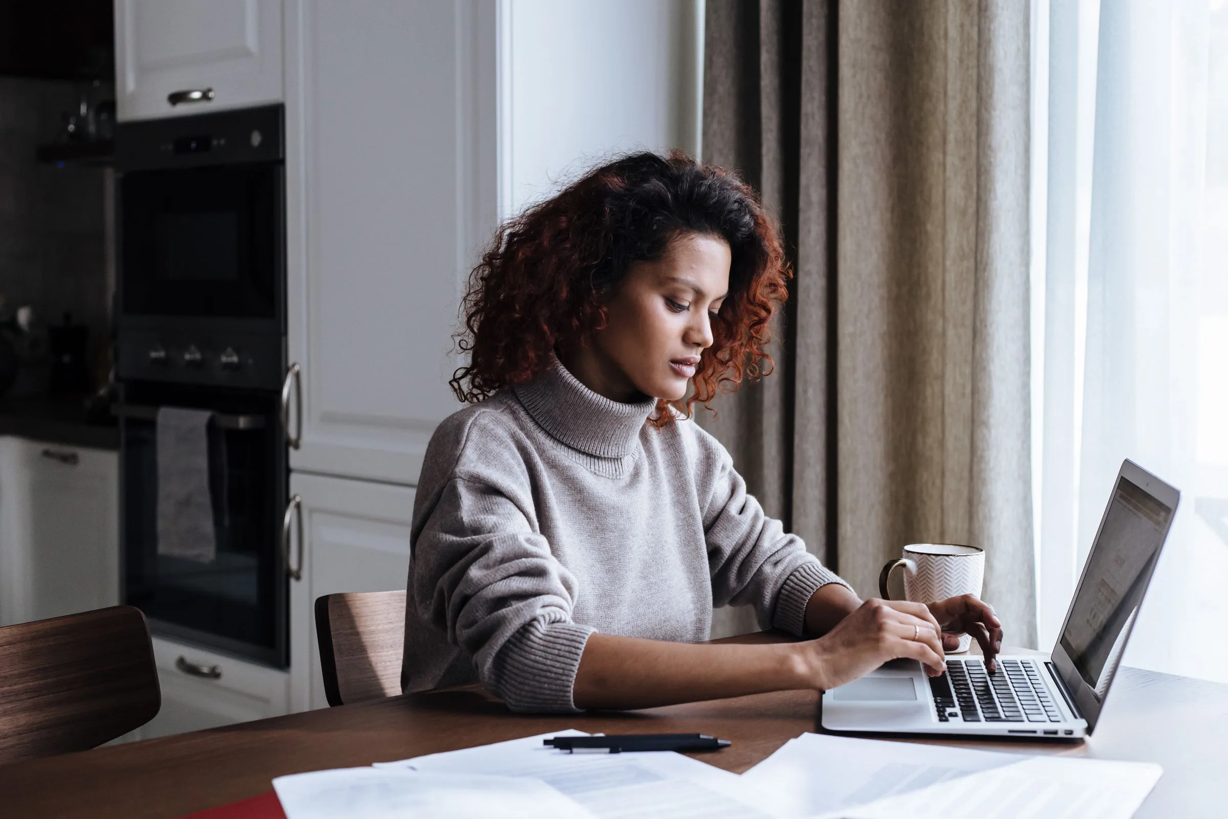 A person at a kitchen table working through a stack of printed property documents with a laptop open beside them