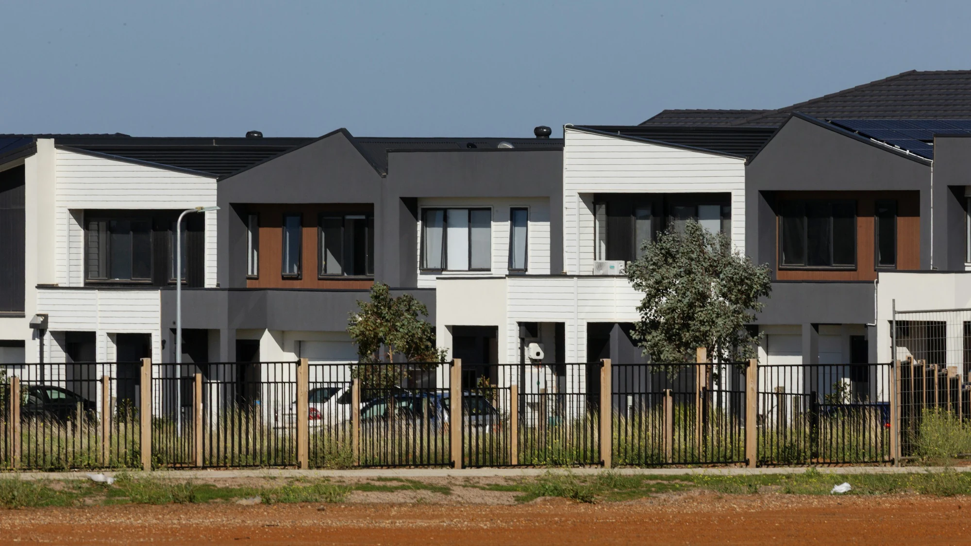 A row of attached two-storey homes with alternating white weatherboard-look and grey rendered walls, each with its own garage and recessed front entry at ground level, behind a black steel fence